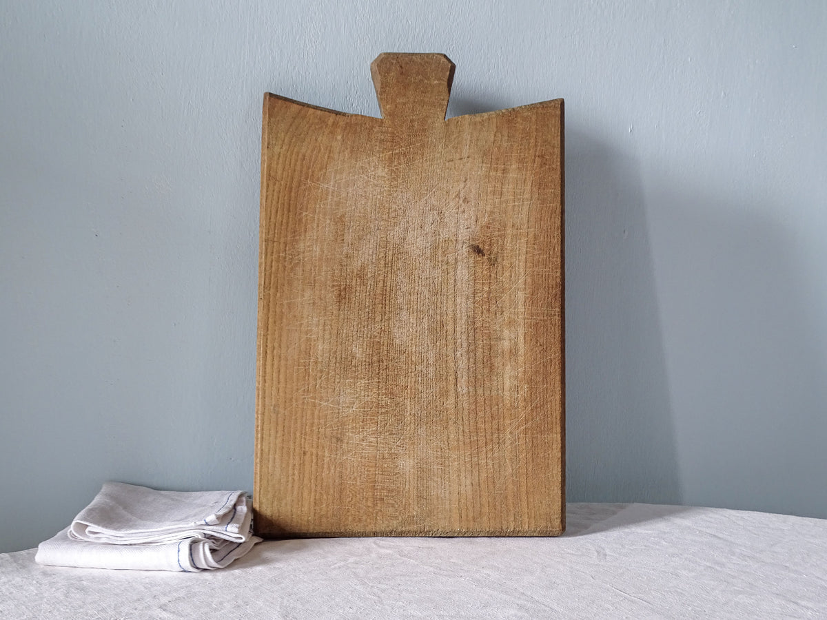 Vintage French wooden cutting board with distinctive flared handle, curved shoulders and warm knife-marked wooden surface.