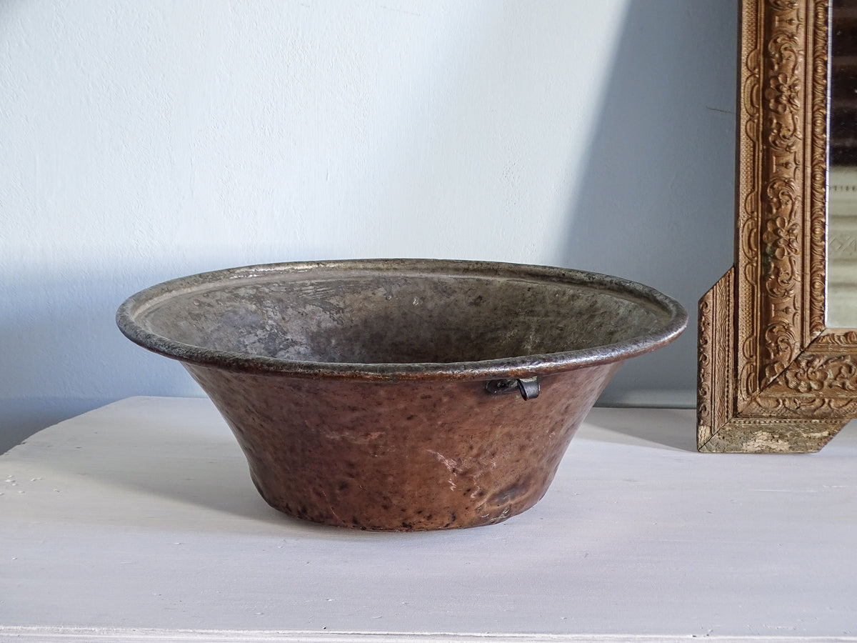 Large antique copper bowl with rustic finish, displayed on a white table next to an ornate gold mirror