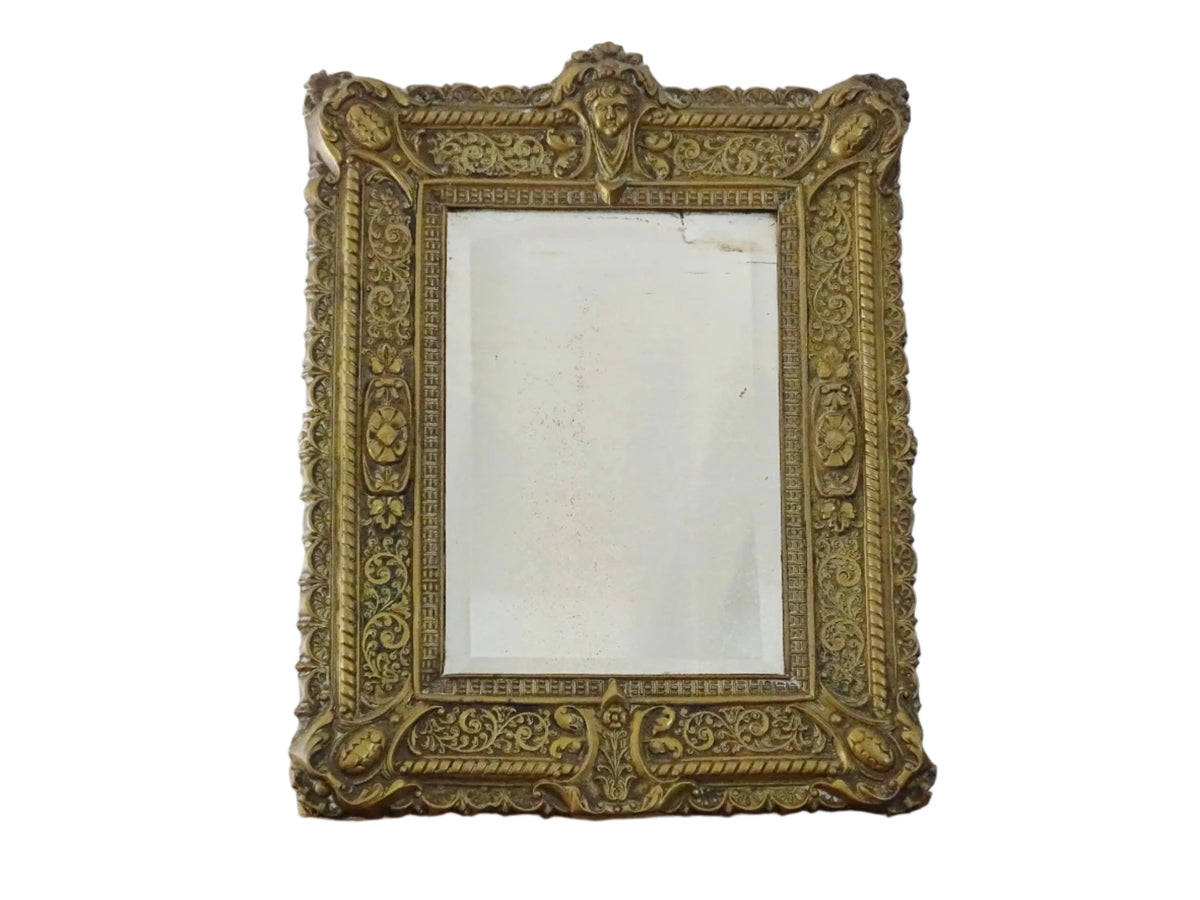 Small French antique brass mirror. The frame is embossed and decorated with a face, flowers and scroll design. Antique French mirror or Foxed mirror.