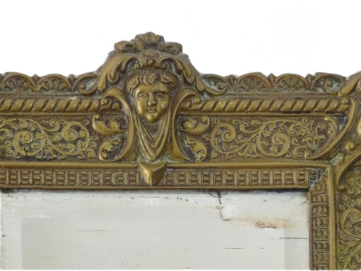 Small French antique brass mirror. The frame is embossed and decorated with a face, flowers and scroll design. Antique French mirror or Foxed mirror.