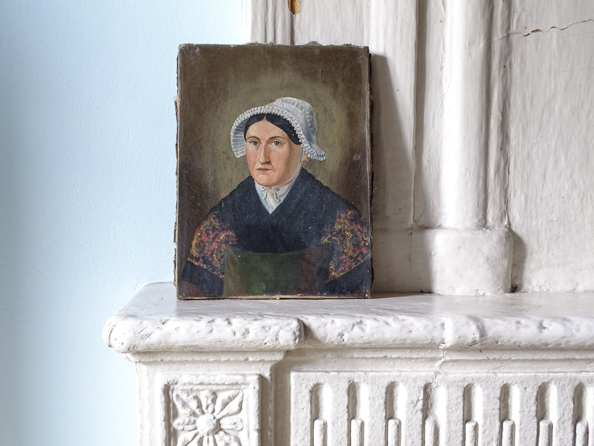 French antique portrait painting of a woman in a white bonnet and dark dress with floral embroidery, displayed on a white stone mantelpiece against a pale blue wall.