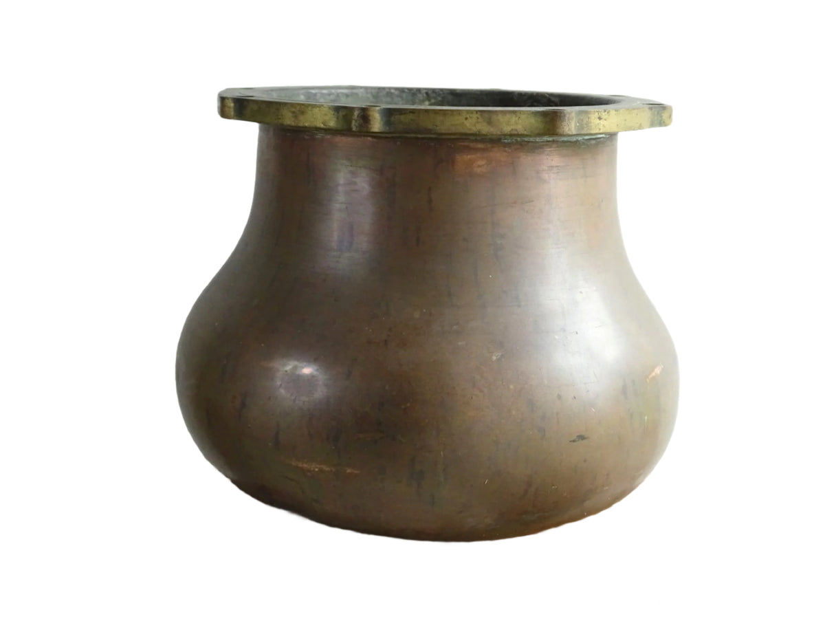 A large antique copper cauldron with a thick brass rim featuring evenly spaced holes, displayed on a rustic wooden surface. The cauldron has a weathered patina, showcasing its aged character.
