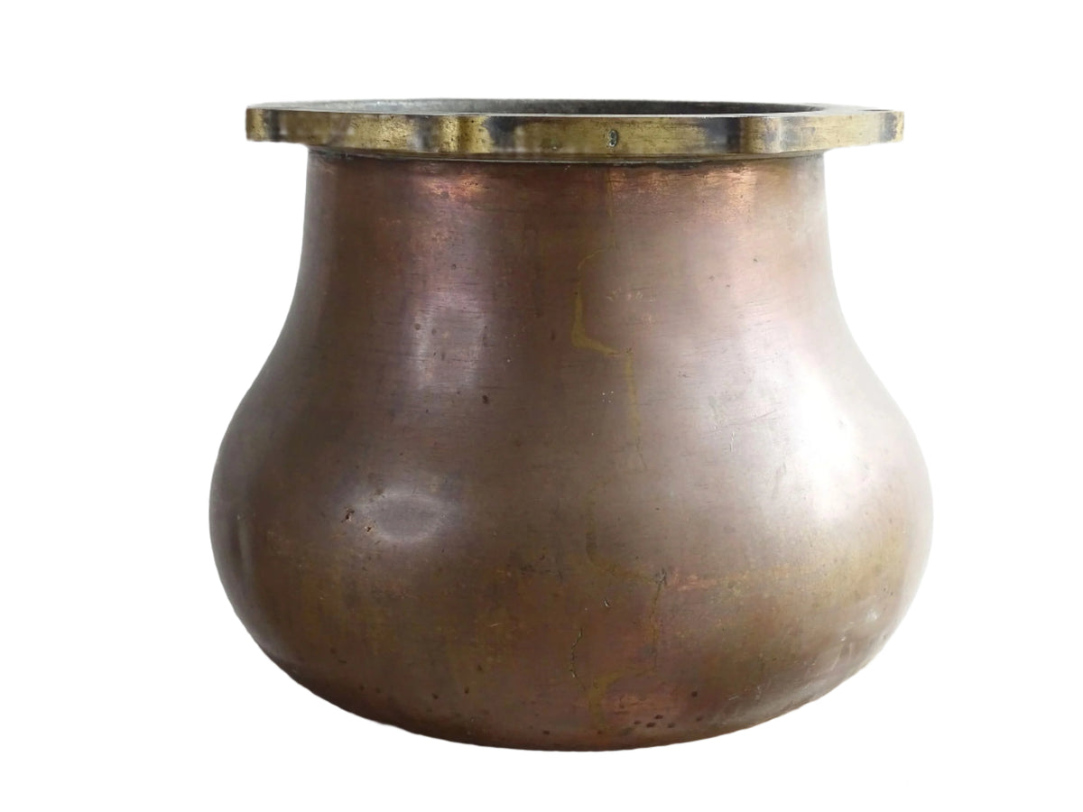 A large antique copper cauldron with a thick brass rim featuring evenly spaced holes, displayed on a rustic wooden surface. The cauldron has a weathered patina, showcasing its aged character.