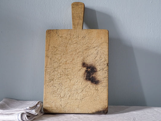 Vintage French wooden cutting board with long handle, pale wood, extensive knife marks and an old scorch mark on one face.