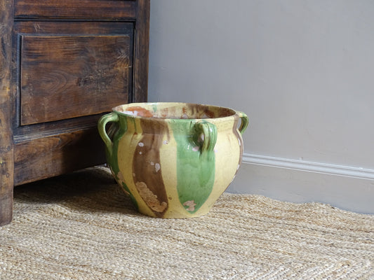 French antique ceramic planter with yellow, brown and green glaze. The glazed terracotta pot sits on a natural jute rug next to an antique wooden seat.