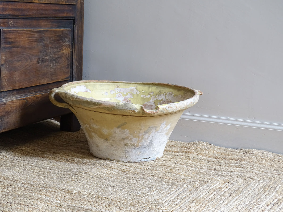 Large French antique confit bowl or tian bowl with yellow glaze interior and unglazed exterior. The ceramic bowl has aged patina and sits on a natural jute rug and next to an antique wooden seat.