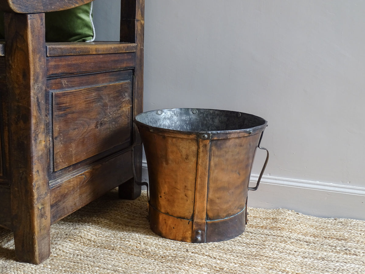 French antique copper water pot or ferrat from the Auvergne region. The copper vessel is sitting on a natural jute jug next to an antique wooden seat.