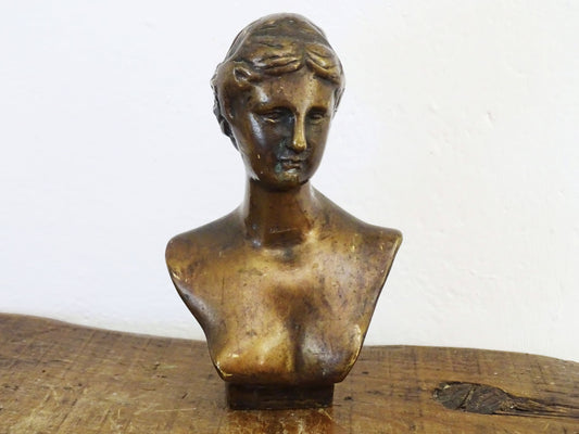 French antique brass bust of a woman. The bust is lightweight and hollow, with finely sculpted facial features and detailed hair. The brass has developed a rich patina, adding character. The form is slightly asymmetrical, with a slanted base.