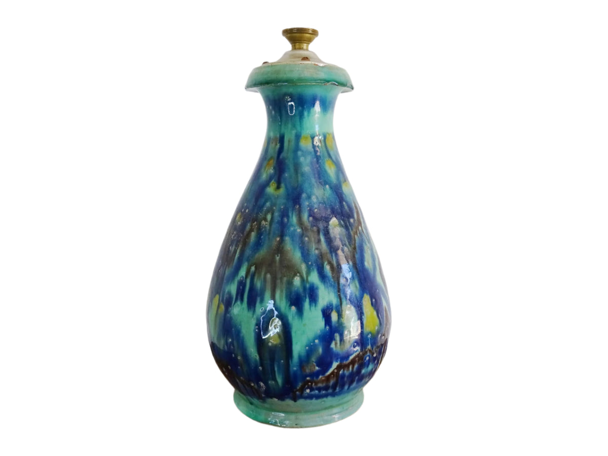 A French vintage ceramic vase lamp base with a bold, multicolored glaze in deep blue, green, and yellow. The organic, dripping pattern covers the surface, giving it a fluid, textured look. The vase has a rounded body that tapers towards a narrower neck, fitted with a brass lamp attachment. It rests on a slightly elevated base.