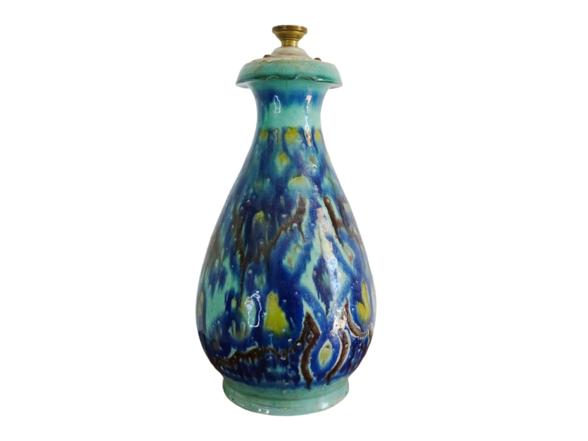 A French vintage ceramic vase lamp base with a bold, multicolored glaze in deep blue, green, and yellow. The organic, dripping pattern covers the surface, giving it a fluid, textured look. The vase has a rounded body that tapers towards a narrower neck, fitted with a brass lamp attachment. It rests on a slightly elevated base.