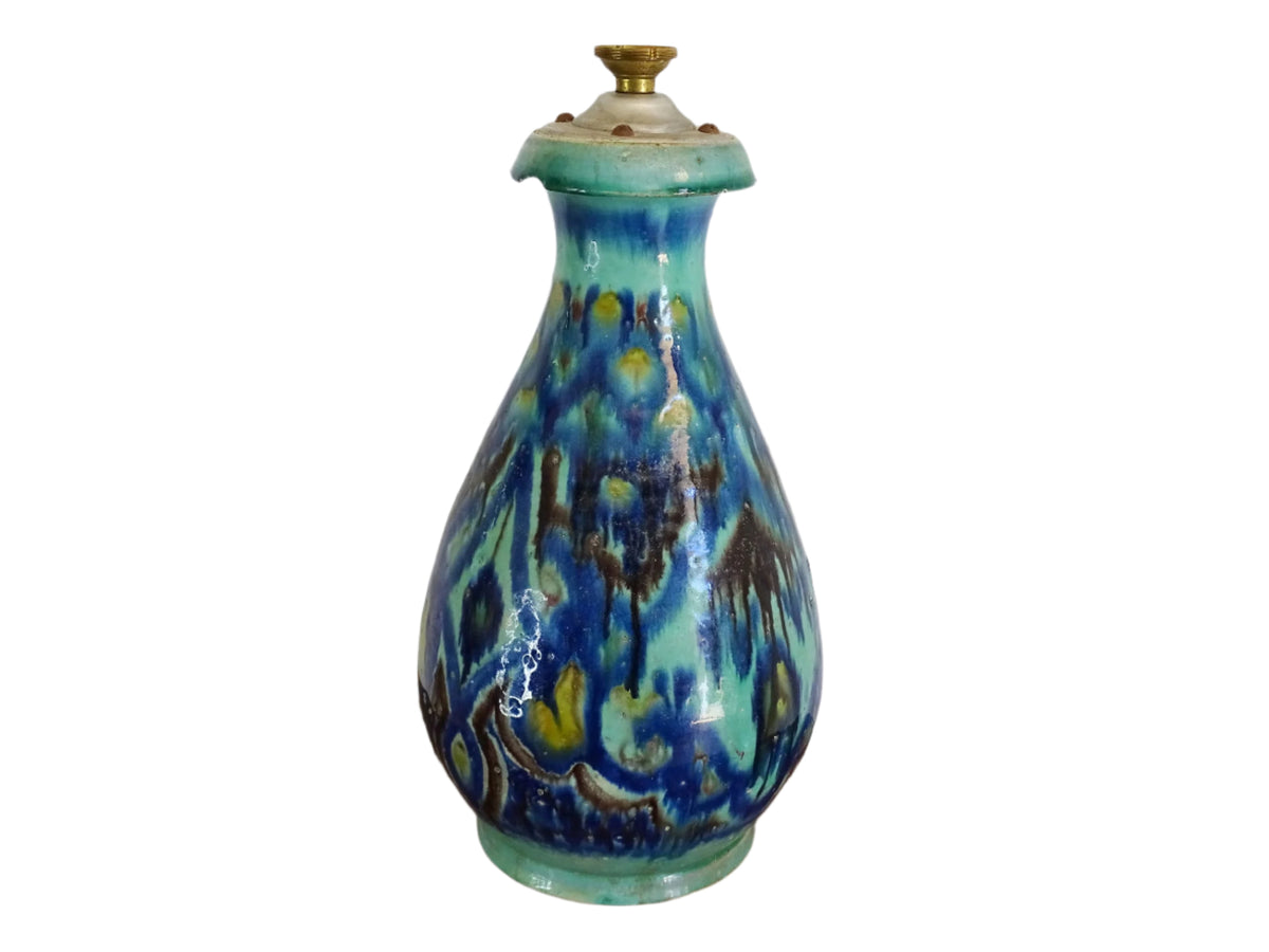A French vintage ceramic vase lamp base with a bold, multicolored glaze in deep blue, green, and yellow. The organic, dripping pattern covers the surface, giving it a fluid, textured look. The vase has a rounded body that tapers towards a narrower neck, fitted with a brass lamp attachment. It rests on a slightly elevated base.
