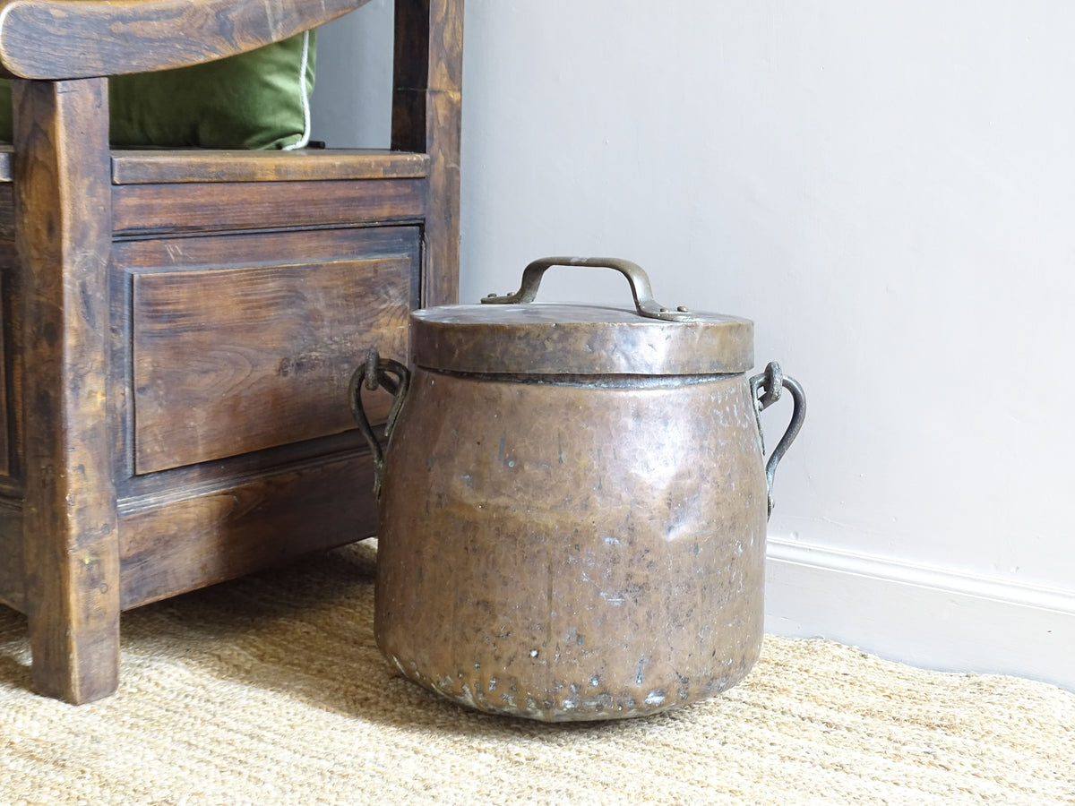 French antique copper cauldron with lid and wrought iron handle from the 18th century. This copper cooking pot with curved base sits on a natural jute, alongside an antique wooden seat.