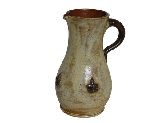 vintage pottery jug with fleur de lys motifs in the style of Roger Guerin. Tall pitcher is glazed in cream, with brown highlights. Display on a sideboard filled with flowers.