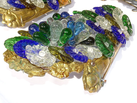 A decorative set of four vintage Murano glass wall lights from Italy. Each sconce features a mix of colourful hand-blown glass pieces in green, blue, amber, clear and deep purple tones, arranged in a raised floral-style pattern. The lights have beautiful ornate gilt metal surrounds with a scrolling form.