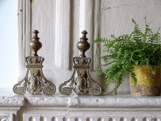 French antique brass firedogs or andirons with scrolling bases and acanthus leaf motifs. Pictured on a country house fireplace mantel next to a yellow confit jar with green fern.