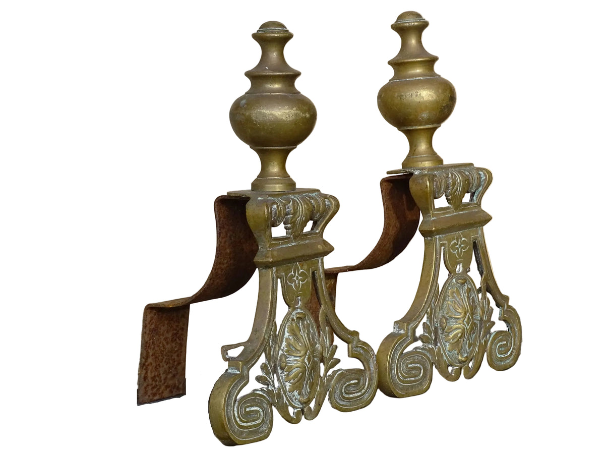 Pair of French antique brass andirons or fire dogs in a Louis XVI revival style. Each andiron is topped with a turned baluster finial. The pierced front plates display an ornate composition of acanthus leaves, scrollwork, and a central oval cartouche framed by laurel branches, typical of neoclassical design.
