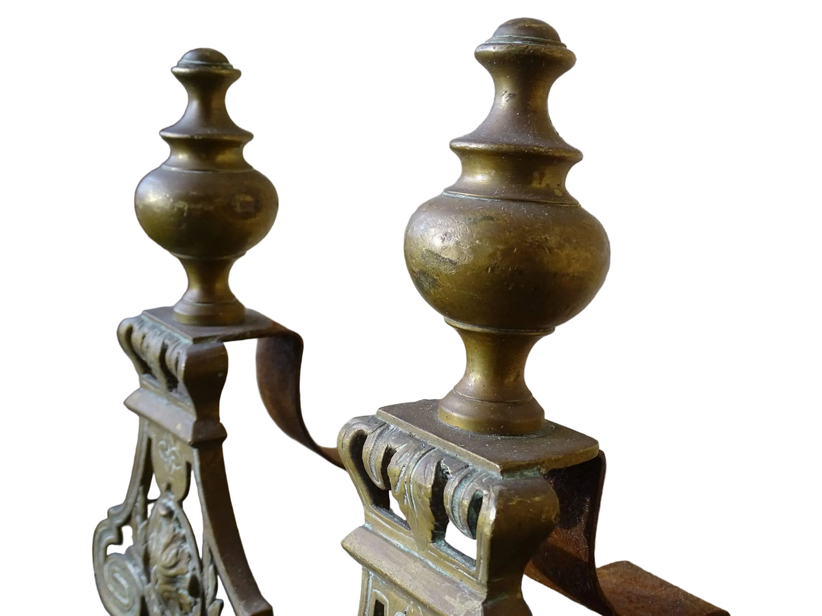 Pair of French antique brass andirons or fire dogs in a Louis XVI revival style. Each andiron is topped with a turned baluster finial. The pierced front plates display an ornate composition of acanthus leaves, scrollwork, and a central oval cartouche framed by laurel branches, typical of neoclassical design.