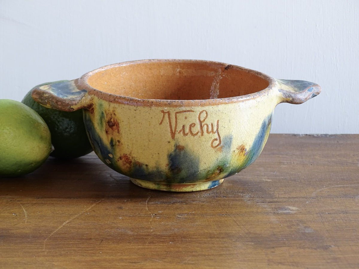 A French vintage ceramic souvenir bowl from Vichy, in the Auvergne region of France. Decorated with hand-applied sponged slip in yellow, green and blue tones. Mid 20th century, with a warm rustic glaze and visible age-related crazing.
