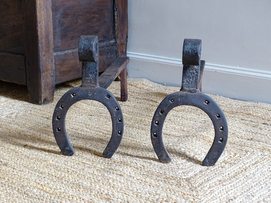 pair of French antique wrought iron andirons or fire dogs, date to the 19th century. They incorporate hand-forged horseshoes, with square-punched nail holes and substantial rear supports. A rustic, agricultural design with authentic forge marks throughout.