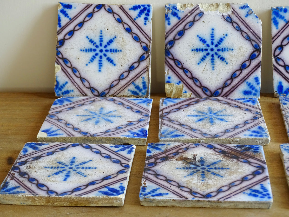 A set of nine French antique Desvres faience tiles in blue & white. These transfer printed, earthenware tiles were manufactured in the late 1900's in Desvres, Northern France. The tiles are back-stamped. Each tile is decorated in cobalt blue on a white tin-glazed ground, centred with a stylised starburst motif and framed within a manganese-purple diamond border.