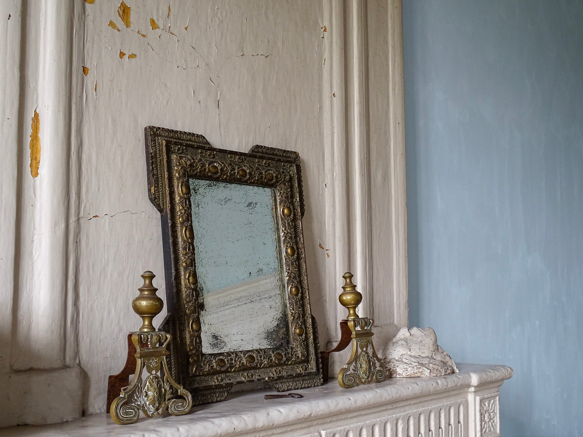 French antique gold mirror with foxed glass on a stone fireplace with brass andirons in a room with pale blue painted walls in a French country house.