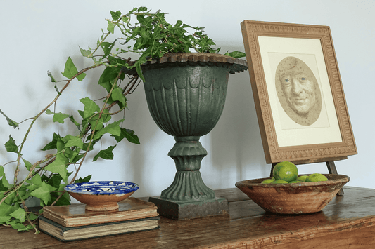 French brocante display with a large green urn, antique pottery bowls, old books, ivy and a framed portrait.