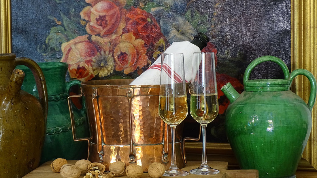 A celebration picture: glasses of sparkling wine, an antique French green glazed pottery gargoulette or jug, an antique French copper ferrat or pot, an antique French walnut oil jug. 