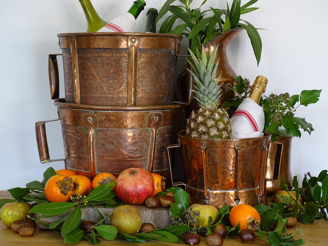 Collection of antique copper ferrats from the Auvergne region of France, displayed in a still life, with fruit, nuts and foliage. The copper vessels are being used as champagne and wine coolers. 