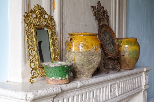 Antique French confit pots, green-glazed pottery and ornate gilt mirror displayed on a weathered white fireplace mantel with framed artwork.