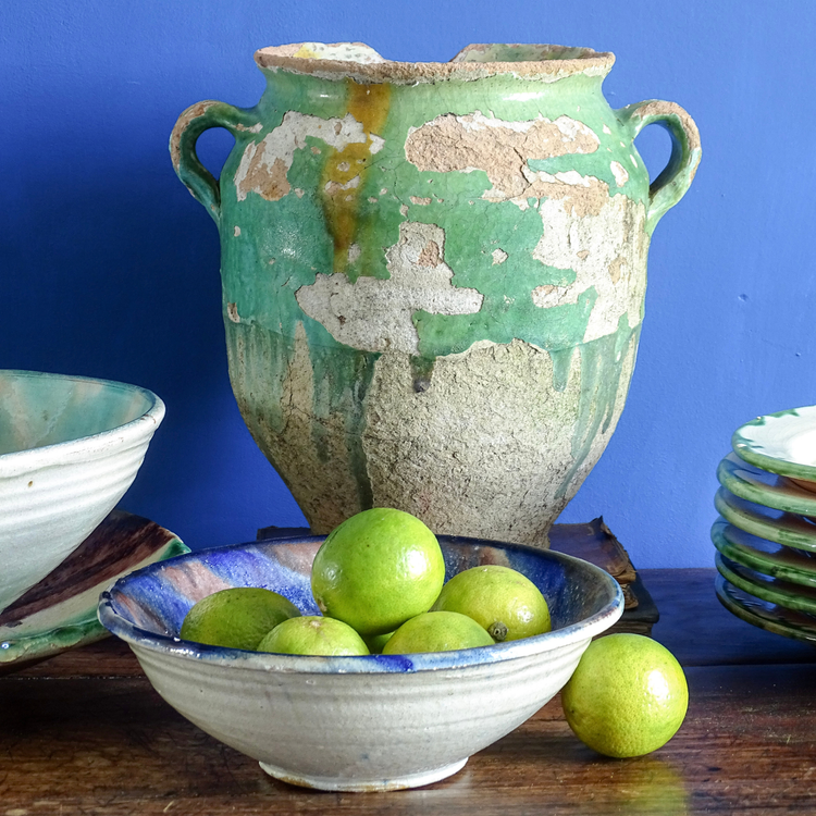 An antique green confit pot with a ceramic bowl filled with limes in front of a blue wall.