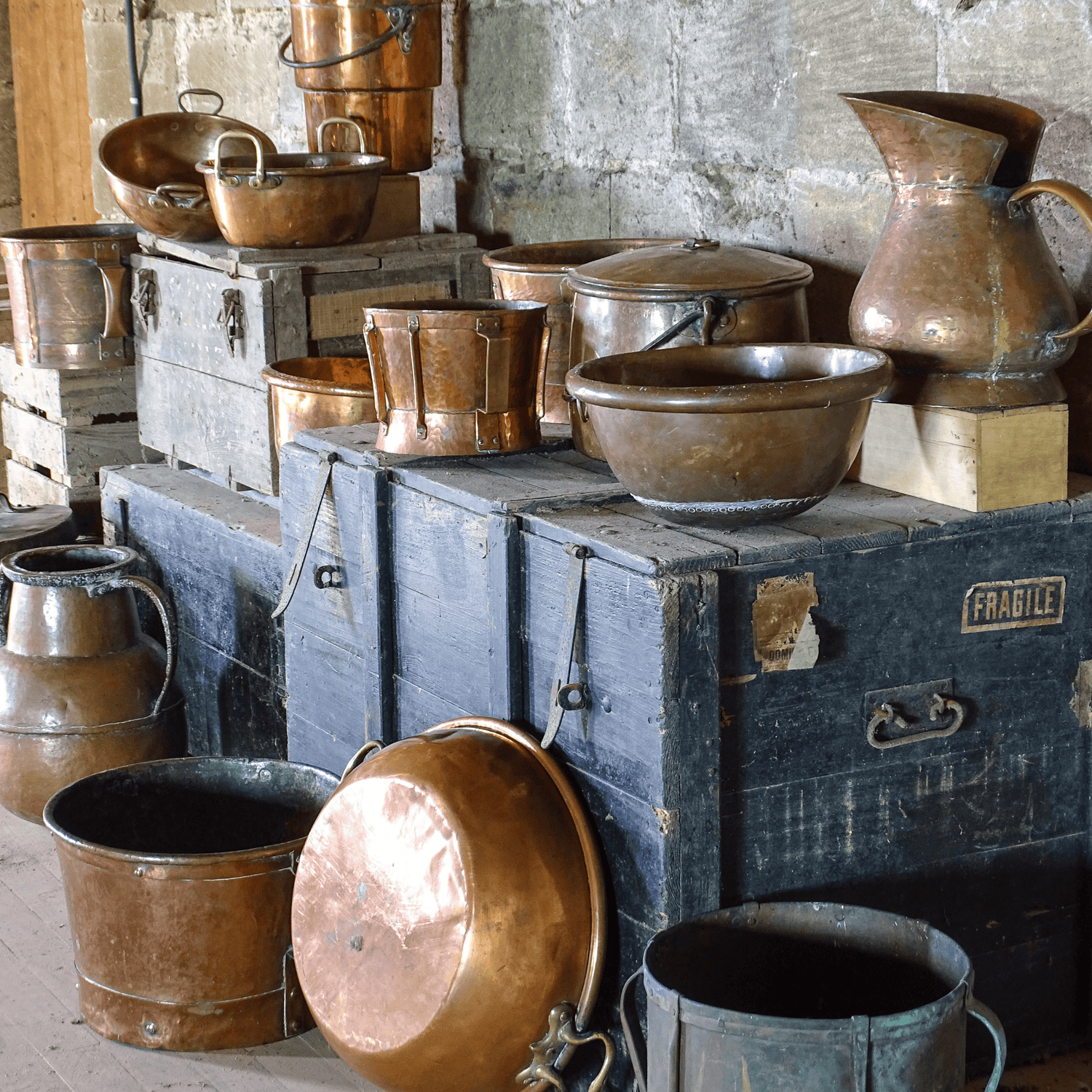 French antique copper pots and vessels displayed on wooden trunks in an attic with stone walls, including basins, pitchers, and cooking pots