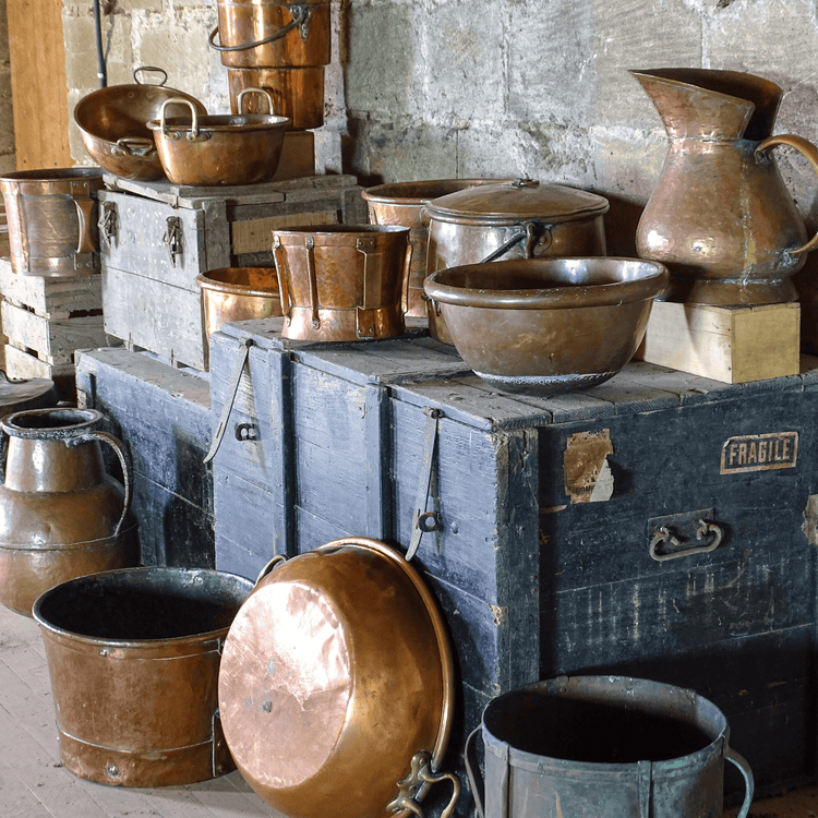 French antique copper pots and vessels displayed on wooden trunks in an attic with stone walls, including basins, pitchers, and cooking pots