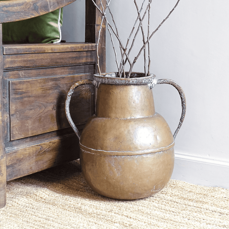 French antique copper vessel with two handles, filled with branches, on a jute rug next to an antique chair