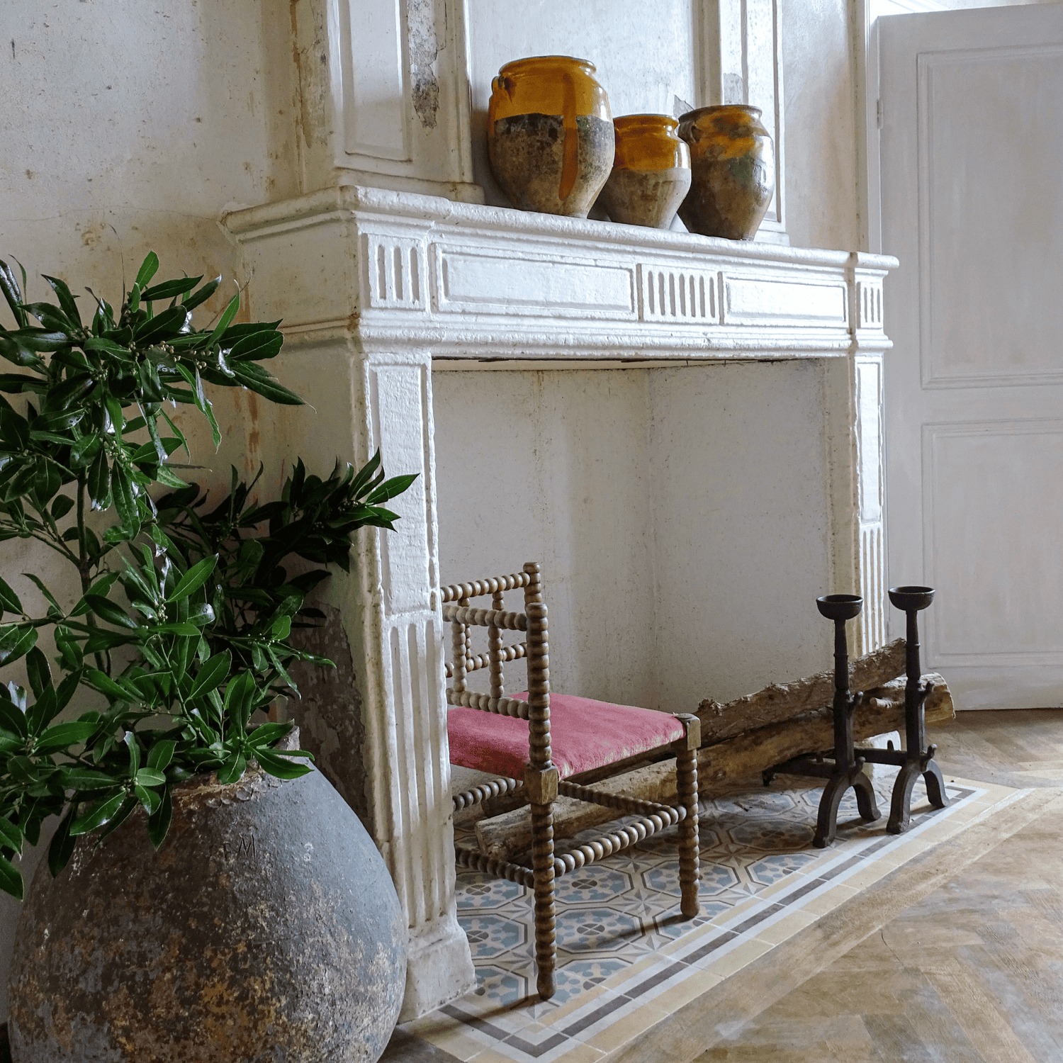 Living room decor, a fireplace with yellow confit pots, a bobbin chair and andirons rest underneath and next to it is a large terracotta vessel or ponne filled with greenery.