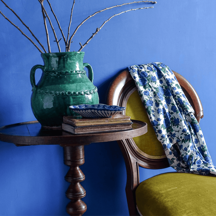 Painted blue walls with a wooden table with a French vintage vase with a green glaze, a small Spanish vintage ceramic Fajalauza dish. Next to the table is a yellow velvet chair draped with a fabric covered in blue roses.