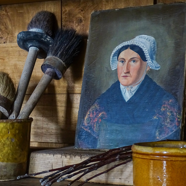 French antique portrait painting of a woman in a bonnet, pictured alongside a collection of old wooden paintbrushes and antique pottery