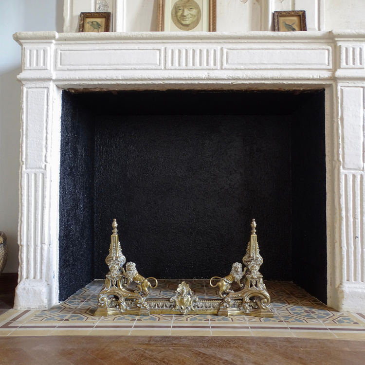 Antique brass firedogs and fireplace fender displayed on a tiled hearth in a traditional French fireplace.
