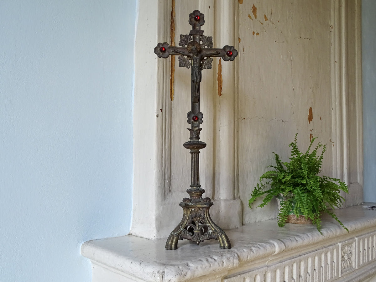 French antique standing cross in gilt metal with red glass stones, pictured with a potted fern on a country house fireplace next to a pale blue wall. 