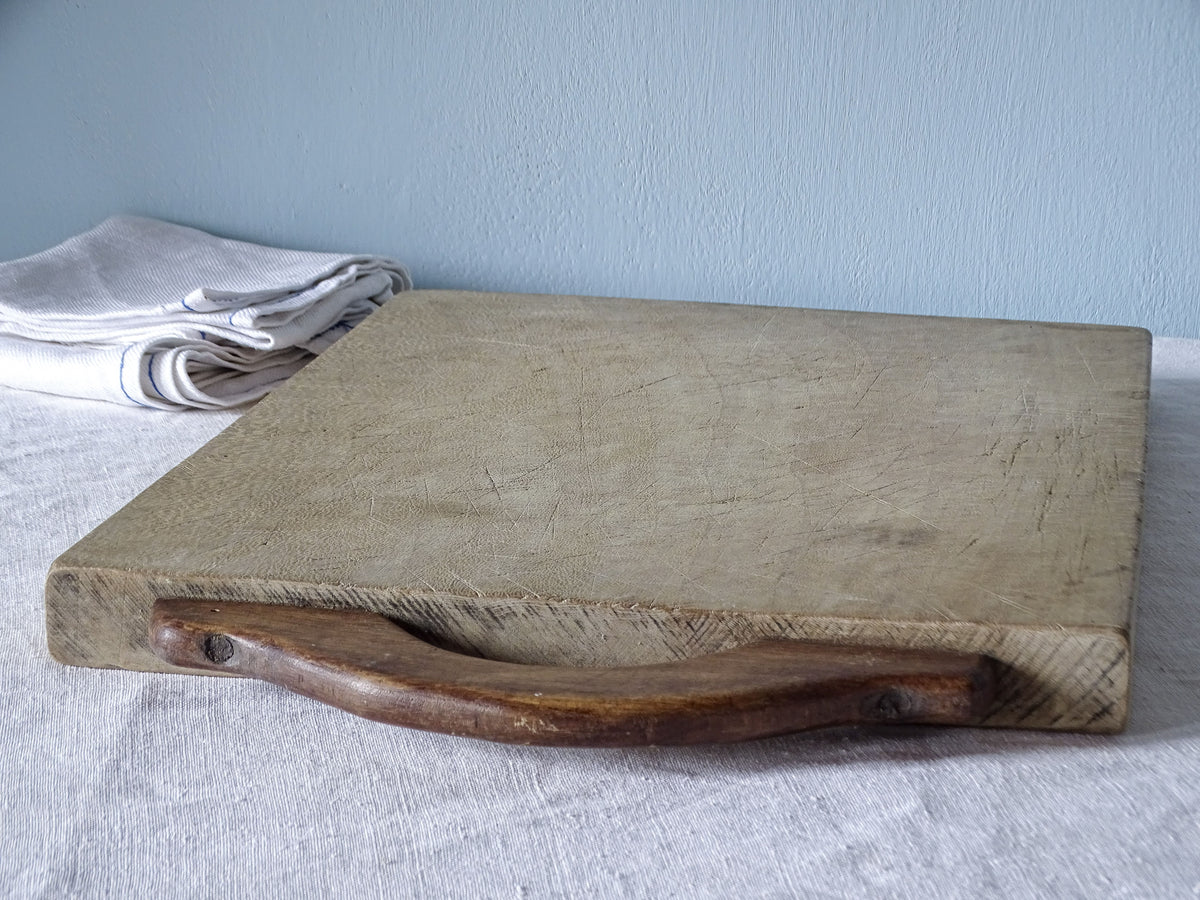 Angled view of vintage French chopping board with a contrasting dark curved wooden handle secured by screws.