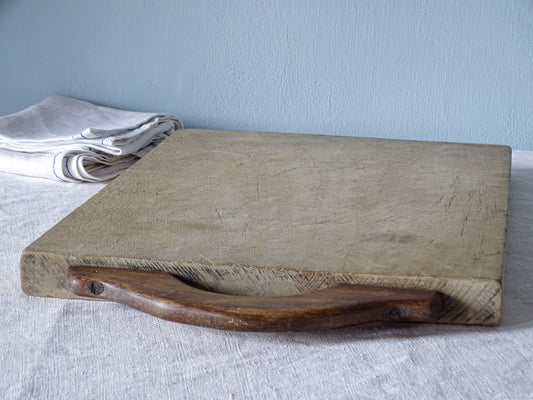 Angled view of vintage French chopping board with a contrasting dark curved wooden handle secured by screws.