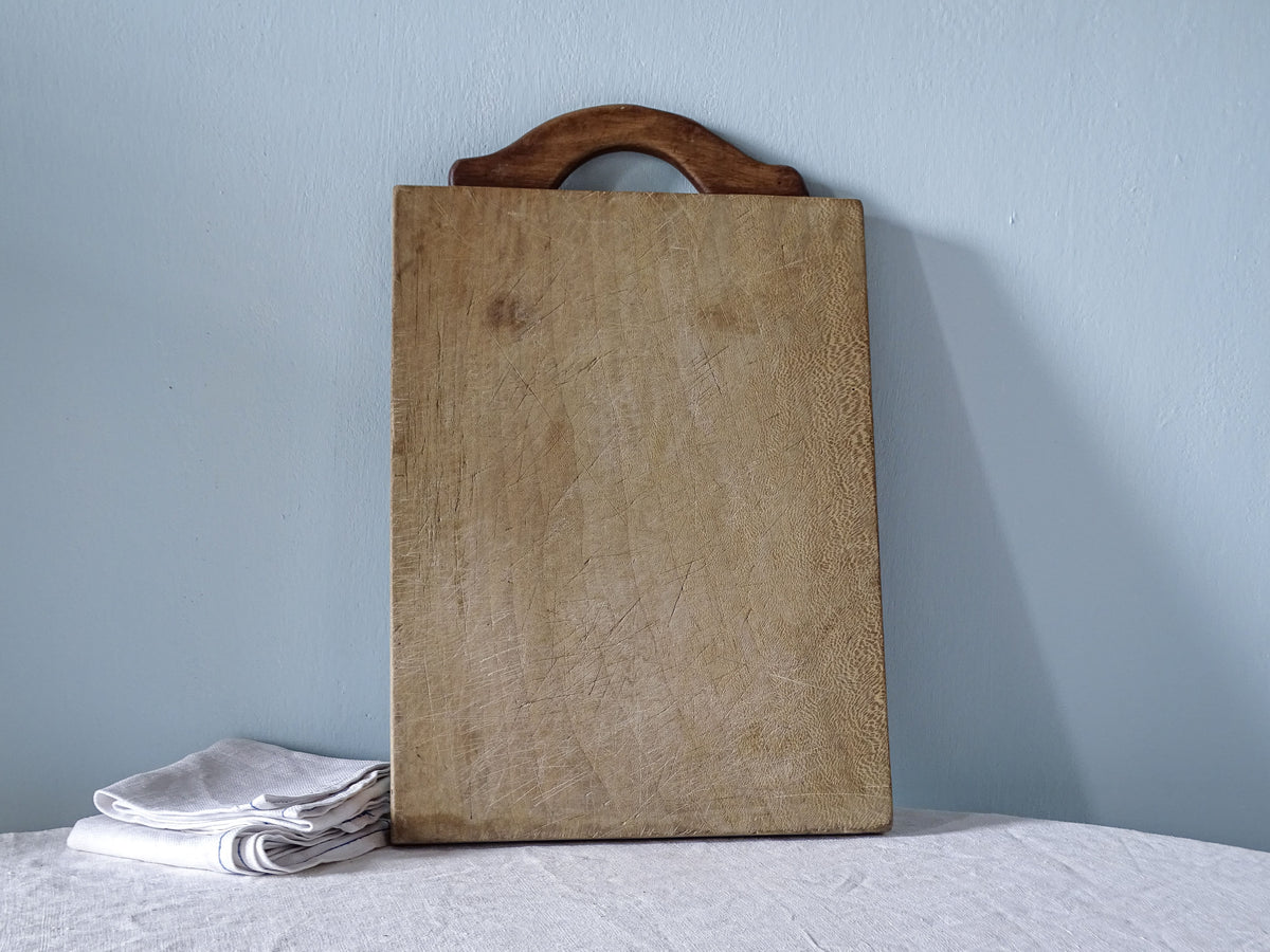 Vintage French wooden cutting board with curved dark wood handle, pale knife-marked surface and rectangular shape.