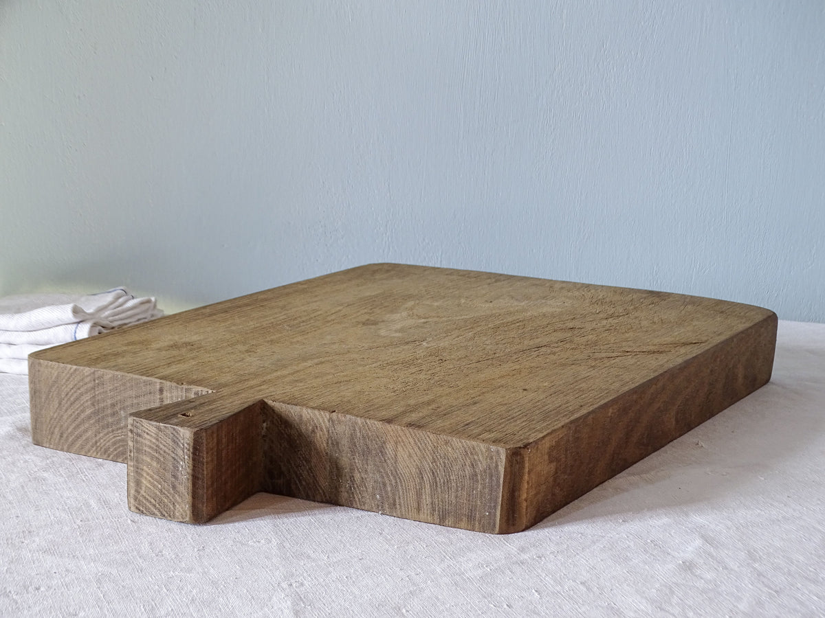 Large antique French wooden chopping board photographed at an angle, showing its rectangular shape, short handle and thick wooden edges.