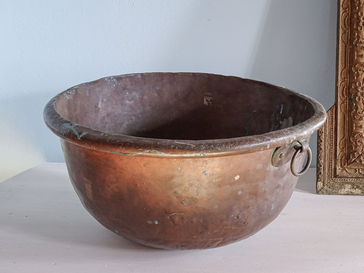 A large French antique copper bowl for confectioners with curved base and loop handle, in front of a pale blue wall