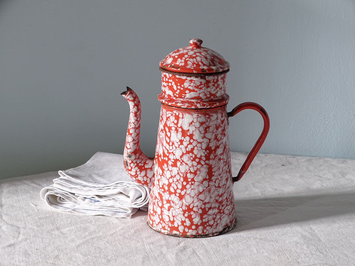 French vintage enamelware coffee pot with a red and white marble pattern, pictured next to antique linen in front of a pale blue wall