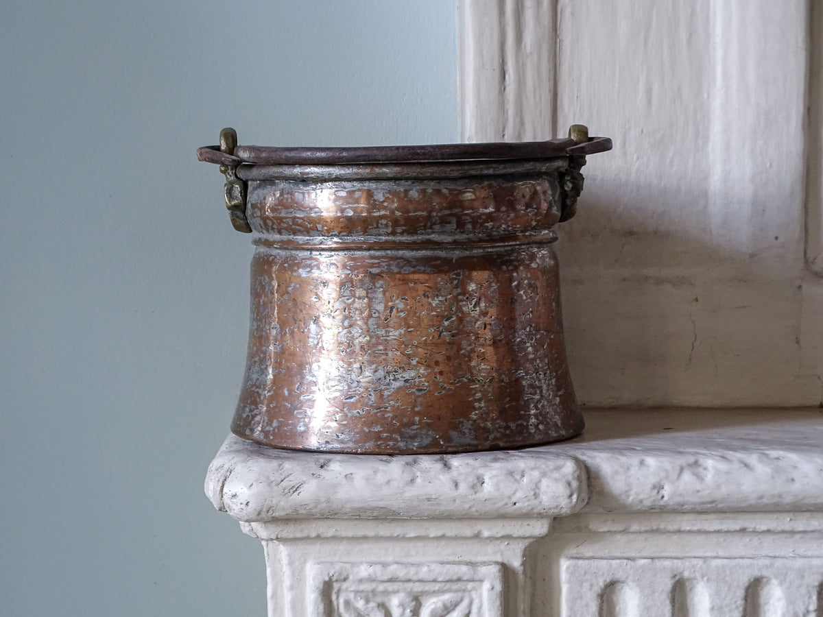French antique copper cauldron with brass handle, displayed on a country house fireplace next to a blue wall