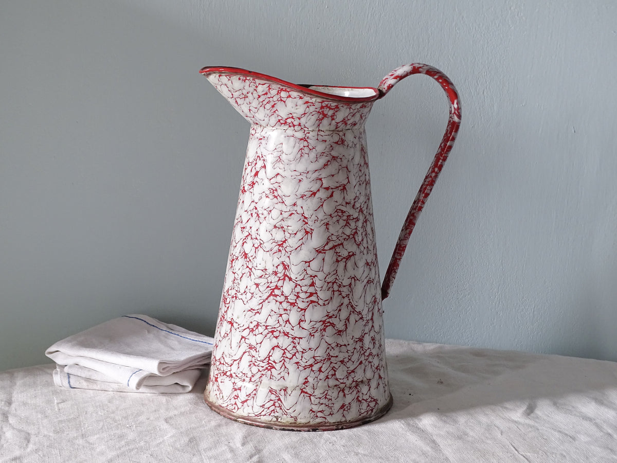 French vintage enamelware pitcher with red marble effect, pictured with antique linen in front of a pale blue wall