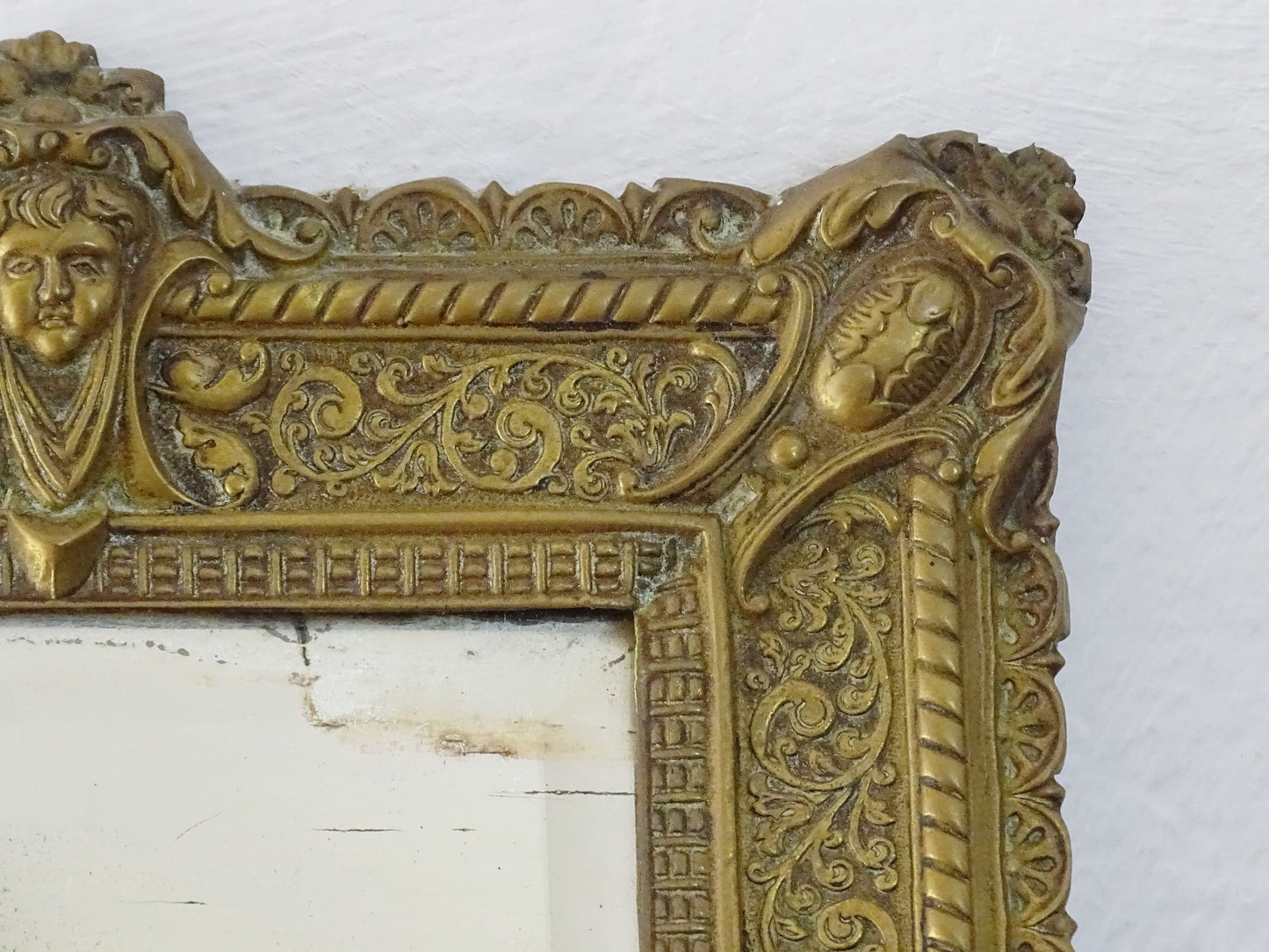 Small French antique brass mirror. The frame is embossed and decorated with a face, flowers and scroll design. Antique French mirror or Foxed mirror.