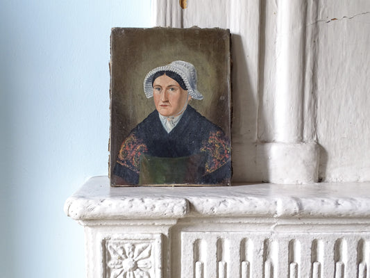 French antique portrait painting of a woman in a white bonnet and dark dress with floral embroidery, displayed on a white stone mantelpiece against a pale blue wall.