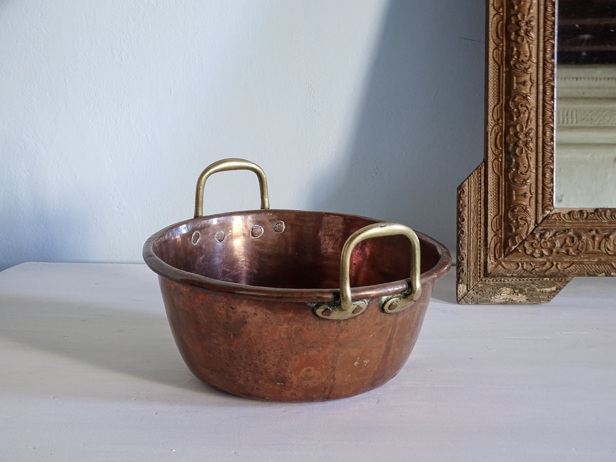 French antique copper confiture pan with two brass handles, sitting on a white surface next to an ornate gold mirror frame against a light blue wall.