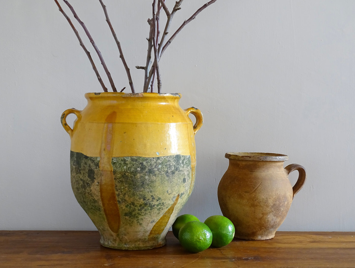 French antique confit pot with yellow glaze filled with branches, alongside a terracotta jug and fresh limes